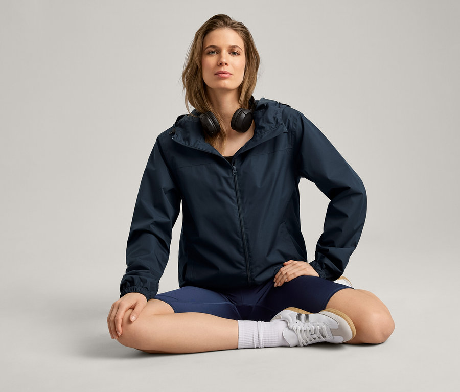 Frau sitzt im Schneidersitz und trägt eine Regenjacke, Kopfhörer, Shorts und Turnschuhe.