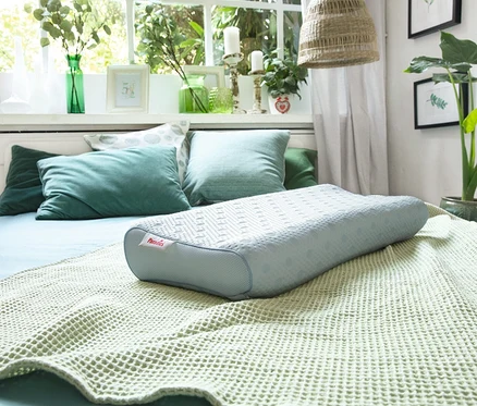 Ein hellblaues Kissen liegt auf einer hellgrünen Decke auf einem Bett mit grünen Kissen vor einem Fenster.