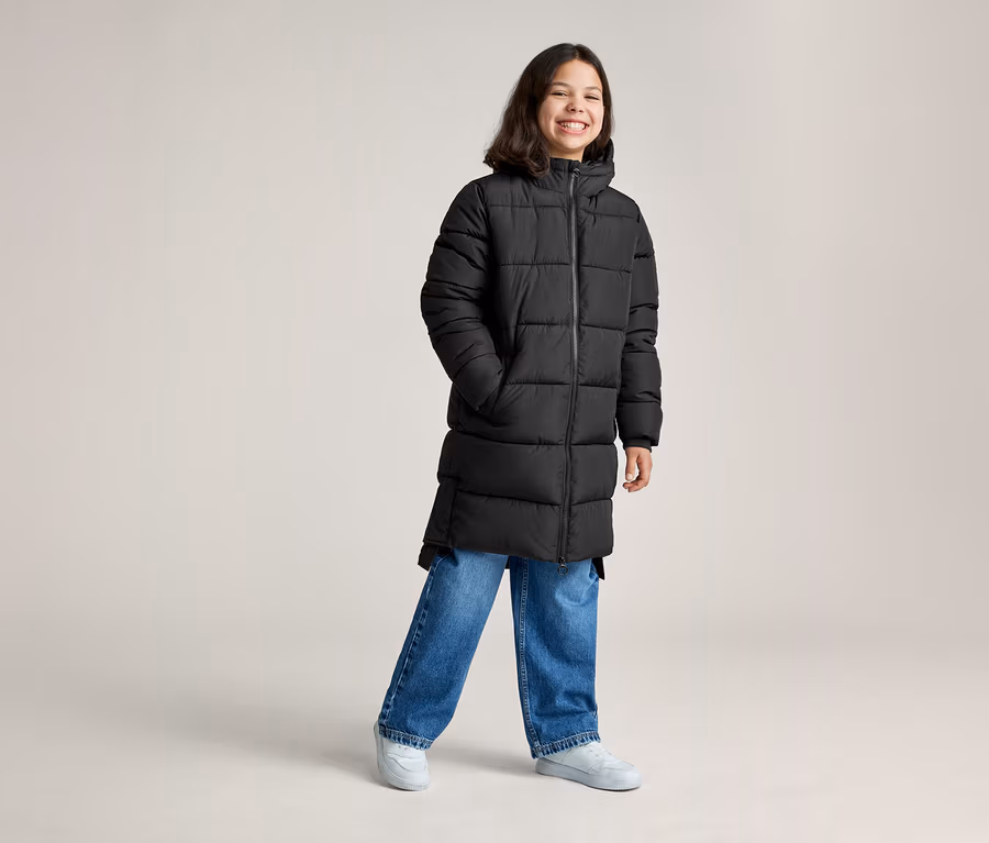 Ein lächelnder junger Mensch trägt einen langen schwarzen Steppmantel, blaue Jeans und hellgraue Sneaker und steht mit den Händen in den Taschen.