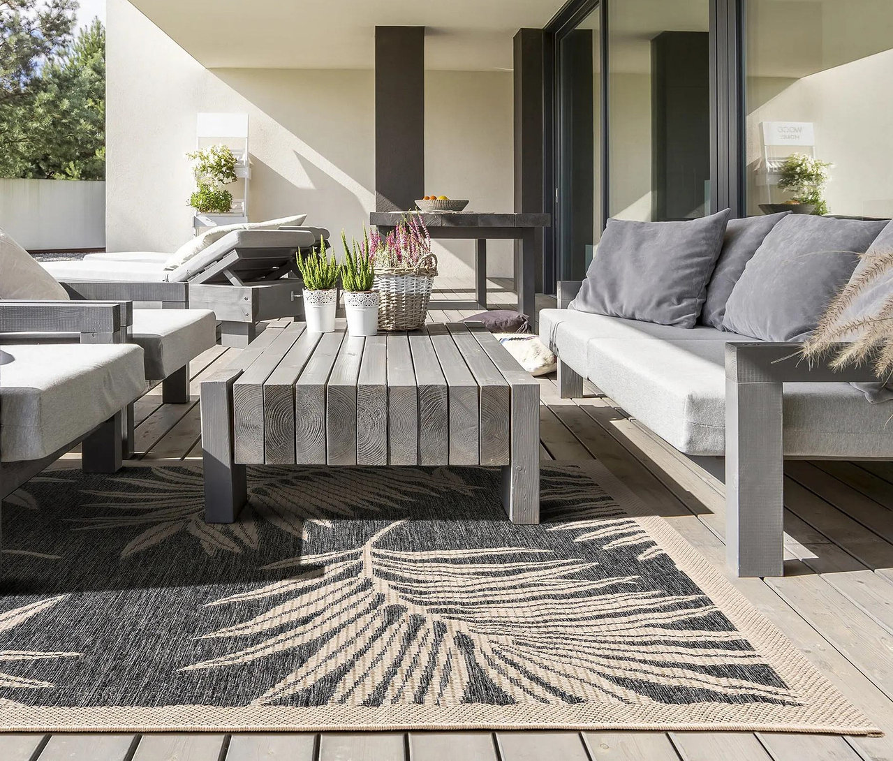 Graue Outdoor-Möbel auf einer Holzterrasse mit einem schwarz-beigen Teppich mit Blattmuster, Pflanzen und einem Esstisch im Hintergrund.