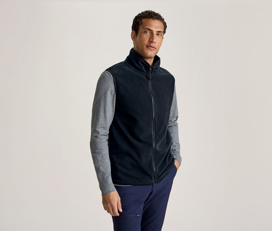 Mann trägt eine schwarze Fleeceweste über einem grauen Langarmshirt und einer dunkelblauen Hose, steht mit einer Hand in der Tasche.