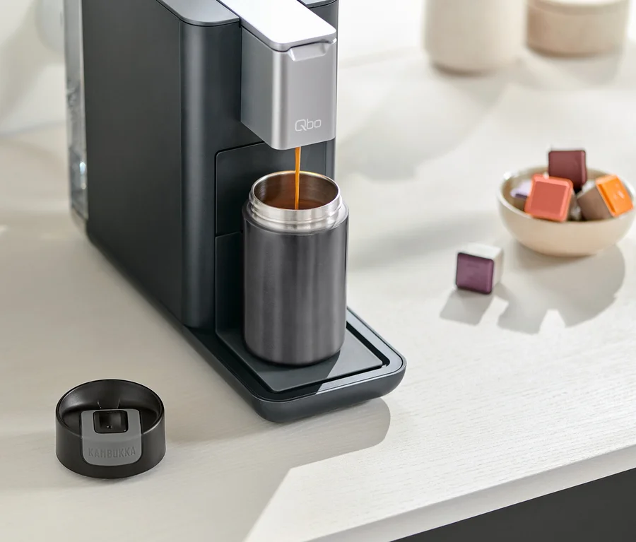 Kaffeemaschine füllt grauen Kambukka To-Go-Becher. Der Deckel des Bechers liegt neben der Kaffeemaschine. Eine Schale mit Kapseln steht daneben.