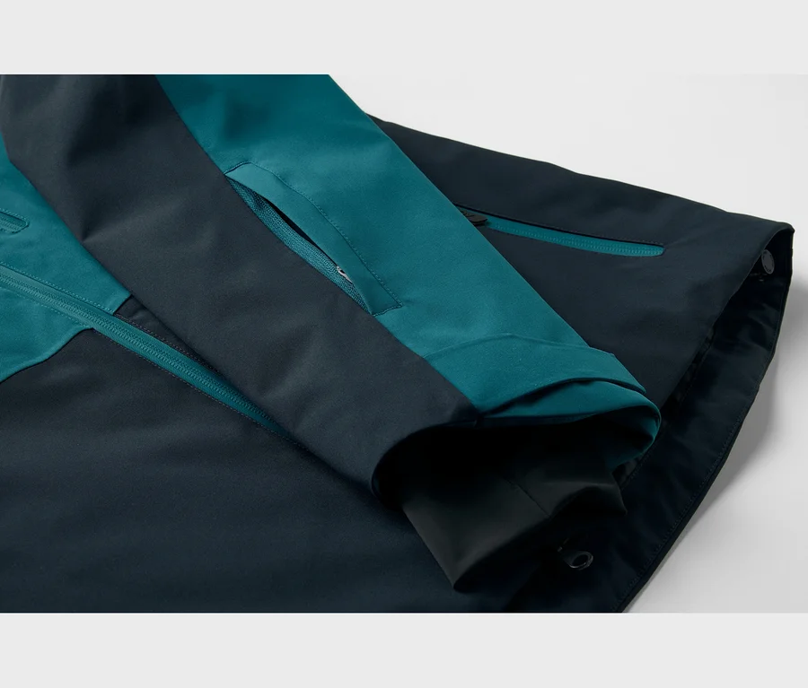 Nahaufnahme einer zweifarbigen Jacke in Dunkeltürkis und Marineblau, mit sichtbaren Reißverschlüssen am Ärmel und am Körper sowie einem verstellbaren Ärmelbündchen.