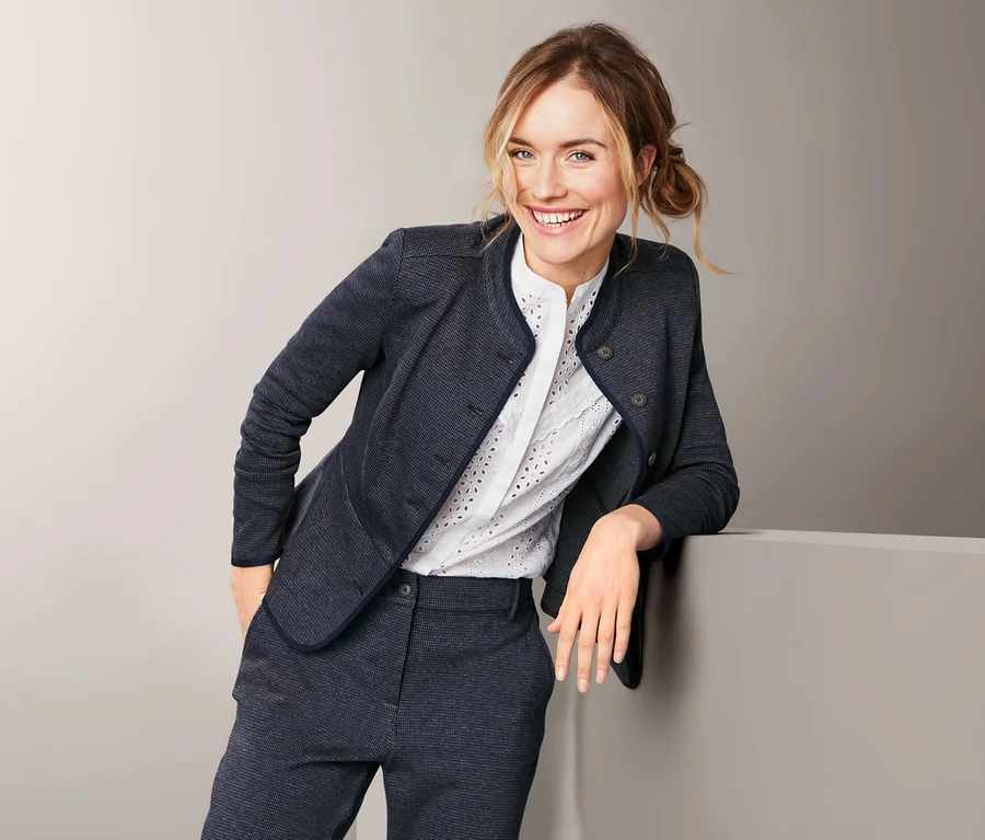 Frau lächelt, an eine Wand gelehnt, trägt einen Sweatblazer und eine Hose.