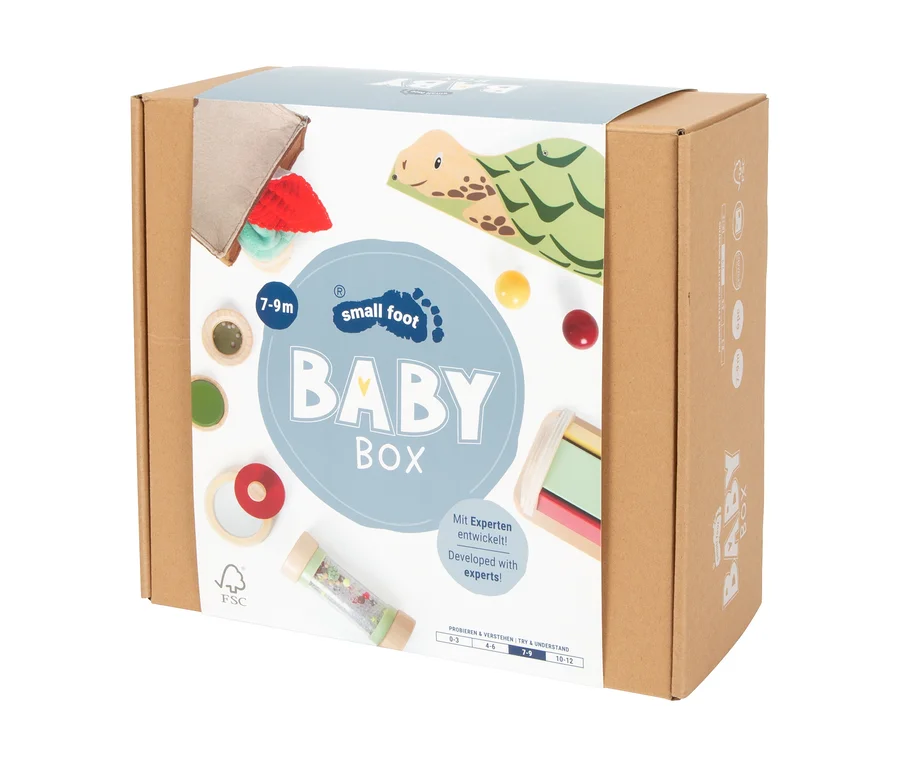 Eine braune Kartonbox der small foot Baby-Box für 7–9 Monate, mit einem Etikett, das verschiedene Babyspielzeuge wie eine Holzschildkröte, Stoffspielzeug, bunte Bälle, Holzringe, einen Rasselstab und ein Holzbausteinspielzeug zeigt.
