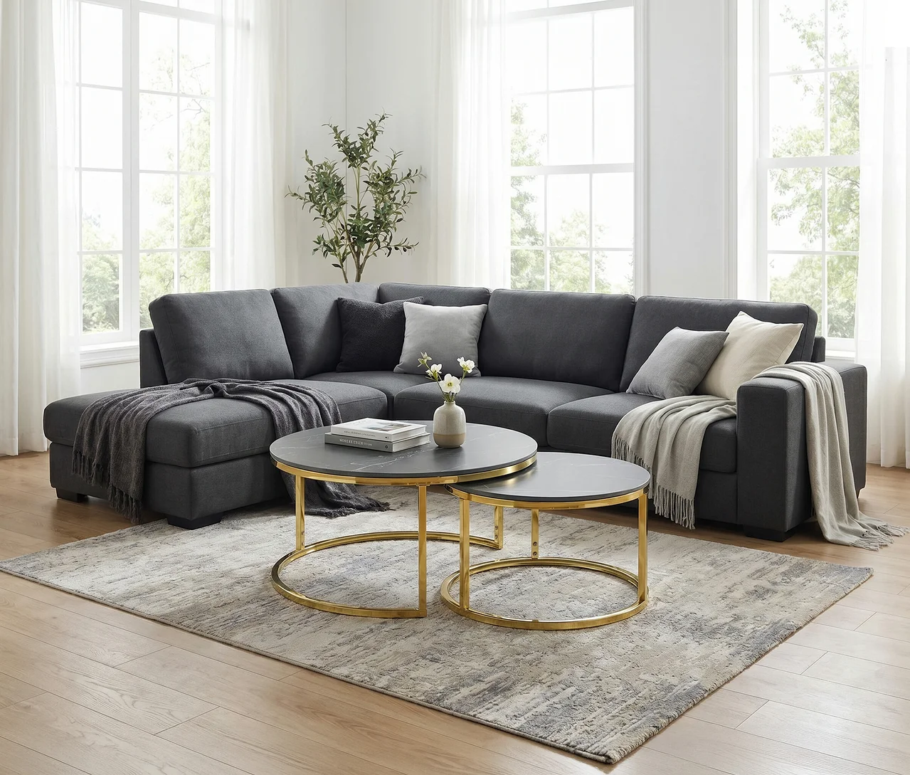 Ein Wohnzimmer mit einem dunkelgrauen L-förmigen Ecksofa, das mit Kissen und Decken dekoriert ist. Davor stehen zwei runde Couchtische mit schwarzer Marmorplatte und goldenen Gestellen auf einem gemusterten Teppich. Im Hintergrund sind große Fenster mit weißen Vorhängen und eine Pflanze zu sehen.