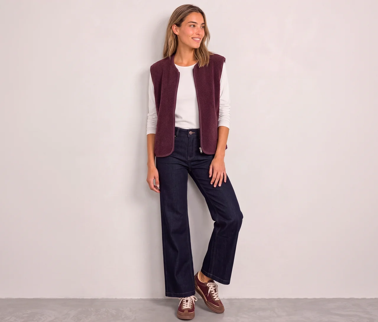 Eine lächelnde Frau trägt eine rote Street One Weste über einem weißen Langarmshirt, dazu weite Jeans und braune Sneaker.