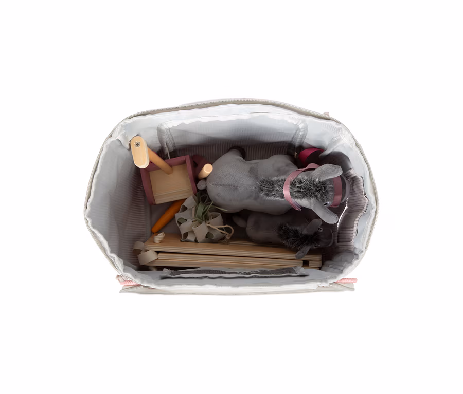 Eine offene hellgraue Tasche mit gestreiftem Futter, gefüllt mit dem small foot Spielset Esel mit Stall. Darin befinden sich zwei graue Plüsch-Esel, Holzteile für einen Stall, eine rosa Holzschubkarre, eine Spielzeugkarotte und eine kleine grüne Pflanze.