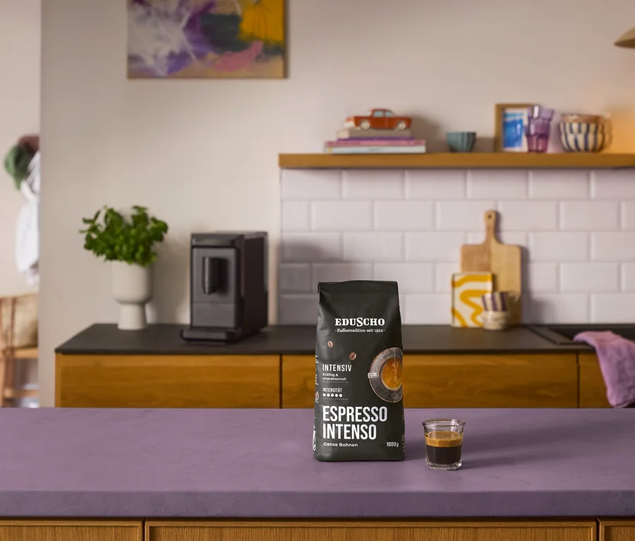 Schwarze Kaffeebohnenpackung "Eduscho Espresso Intenso - 1 kg Ganze Bohne" und ein Espresso auf lila Theke, dahinter ein schwarzer »Esperto mini« Kaffeevollautomat.