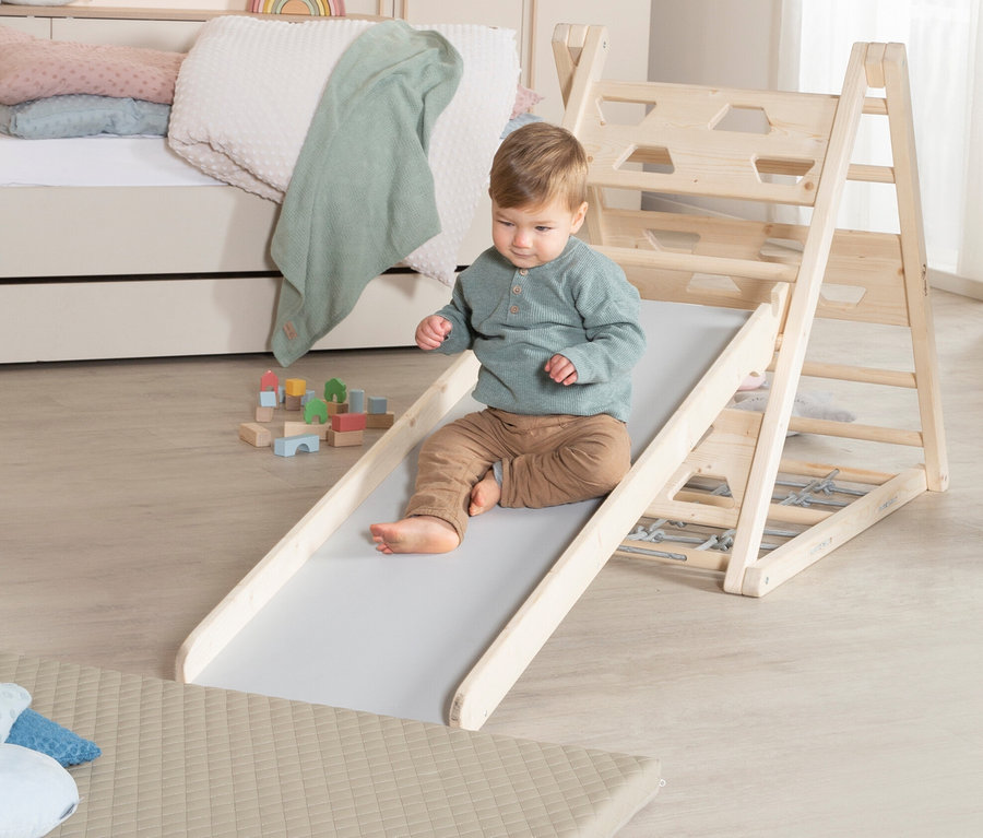 Ein Kind rutscht die Rutsche des roba Kletterdreiecks hinunter, im Hintergrund Spielzeug und ein Bett.