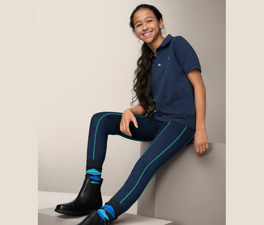 Mädchen sitzt und trägt eine Reittight mit recyceltem Material, ein blaues Reitpolo und schwarze Stiefel.