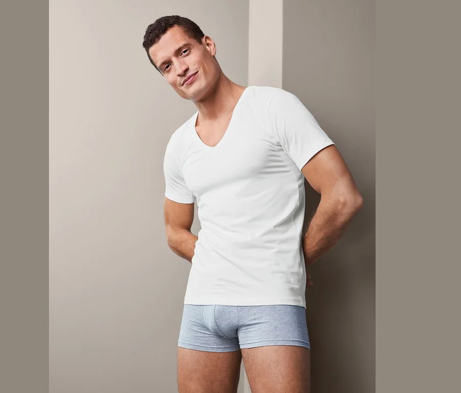 Mann posiert in einem weißen Funktionsunterhemd und grauen Boxershorts. Zu sehen sind 2 Funktionsunterhemden.