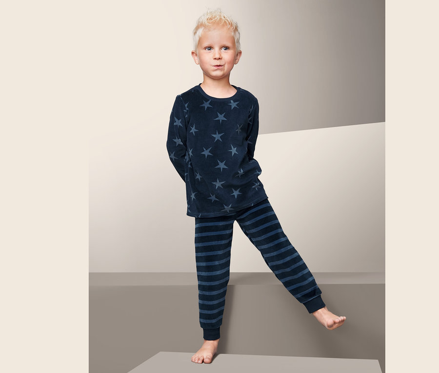 Ein Junge steht barfuß auf einem grauen Würfel und trägt einen Nicki-Pyjama mit Sternen und Streifen.