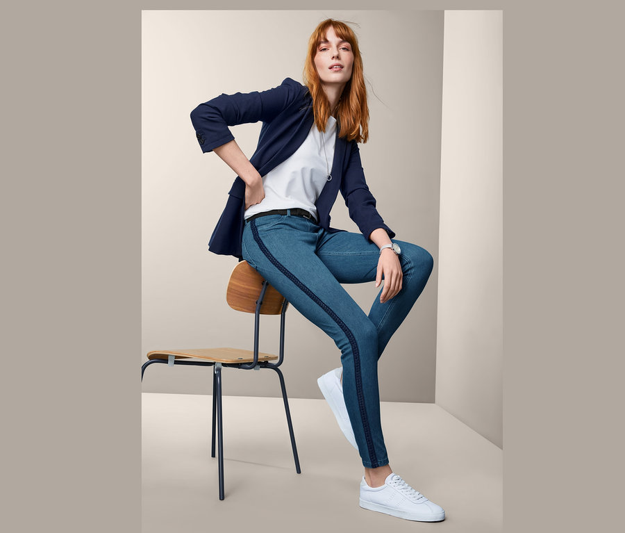 Eine Frau sitzt auf einem Stuhl und trägt blaue Jeggings, ein weißes T-Shirt, einen blauen Blazer und weiße Turnschuhe.