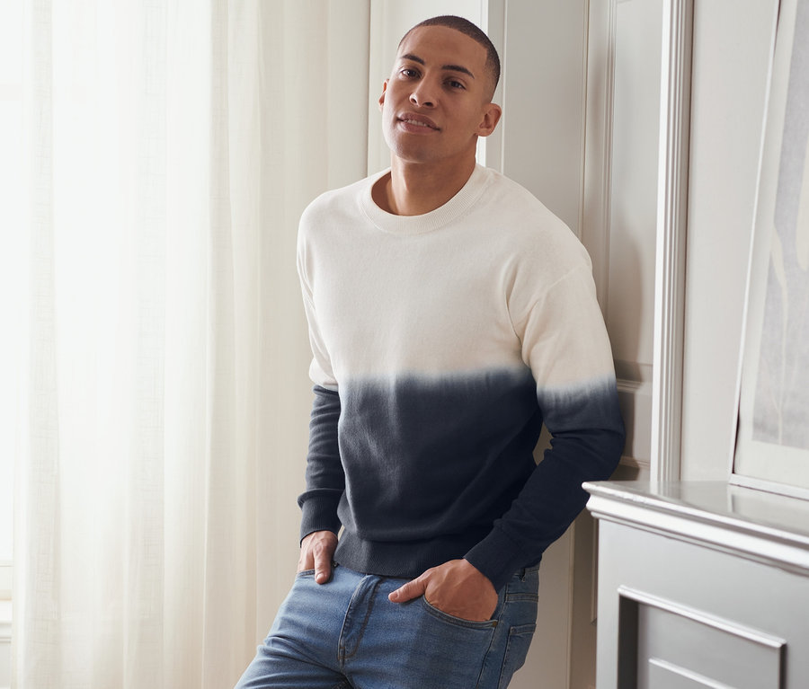 Mann lehnt an Wand, trägt Dip-Dye Pullover, Jeans und Turnschuhe.