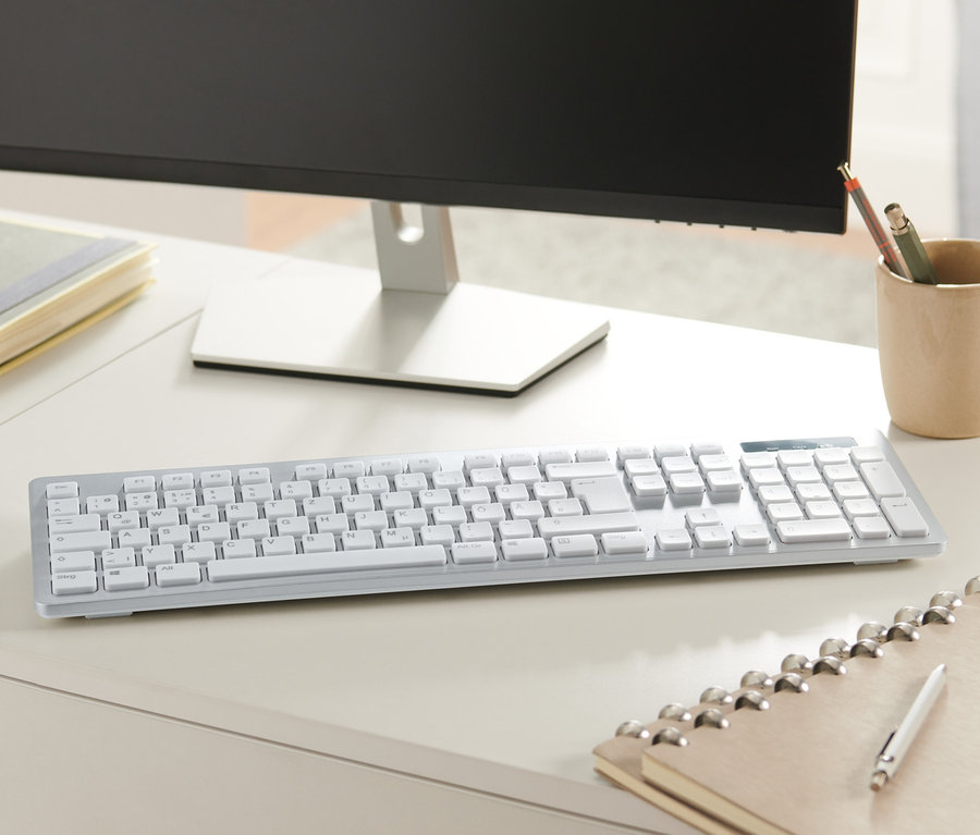 Eine silberne Tastatur liegt auf einem weißen Schreibtisch neben einem Monitor, einem Notizbuch und einem Stiftehalter.