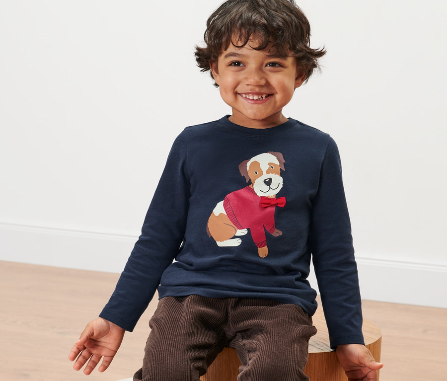 Ein lächelnder Junge sitzt und trägt ein Kleinkinder-Langarmshirt mit einem Hundemotiv.