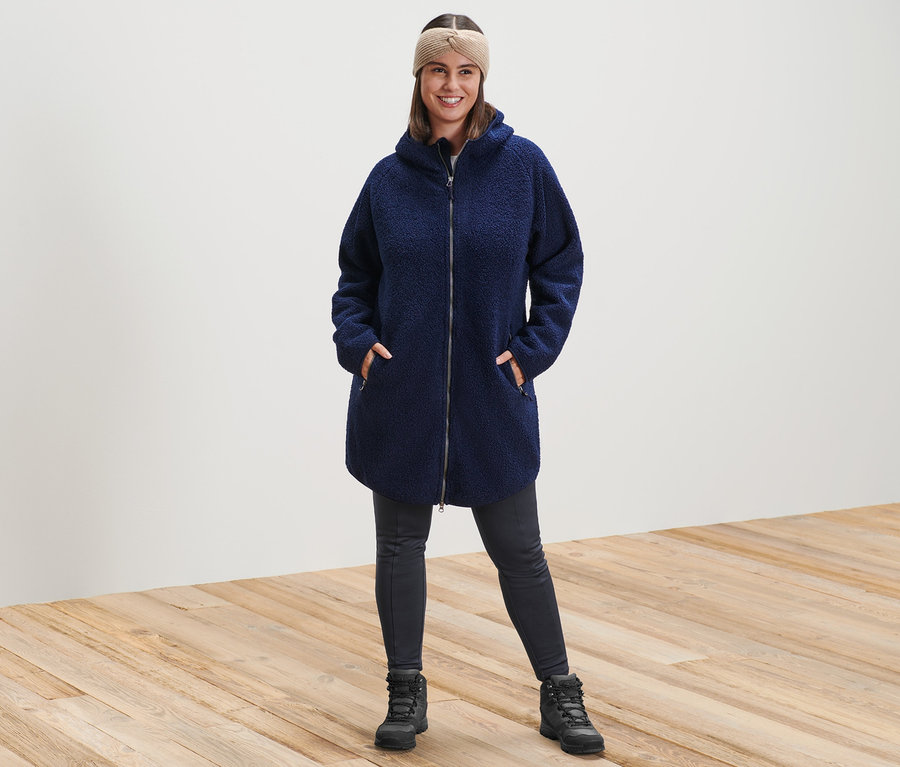 Eine Frau steht und trägt einen Kapuzen-Teddy-Mantel, ein sandfarbenes Strick-Stirnband und eine Thermotight.