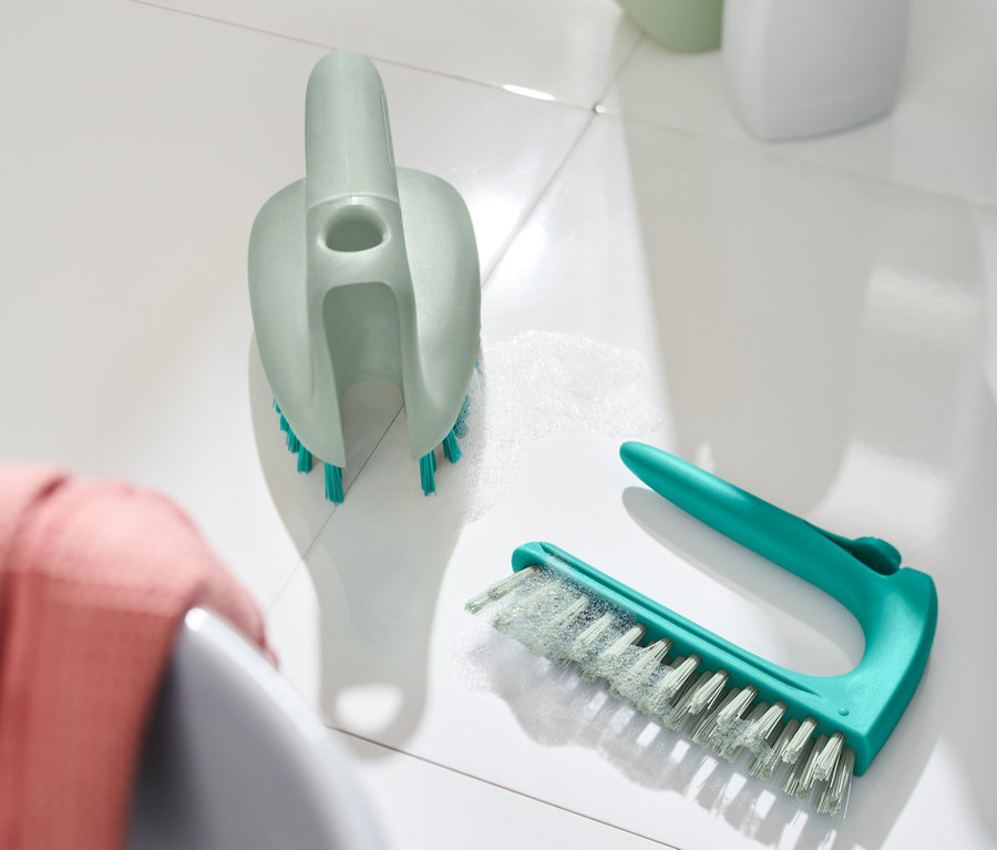 Zwei 2-in-1-Fliesen-Bodenbürsten liegen auf weißen Fliesen neben einem Waschbecken.