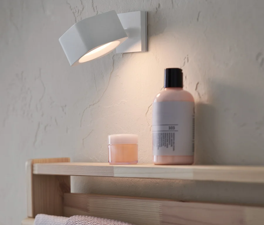 Eine LED-Akku-Wandleuchte ist an der Wand montiert. Auf einem Regal darunter stehen Kosmetikartikel.