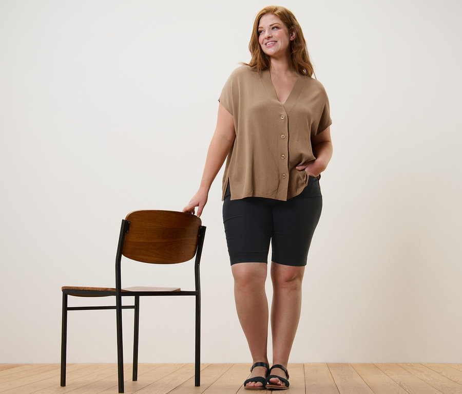 Eine Frau steht neben einem Stuhl und trägt einen beigen, kurzärmligen Cardigan und Bengalin-Bermudashorts.