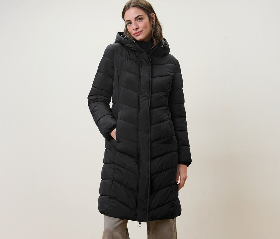 Frau posiert in einem schwarzen Steppmantel mit Kapuze.