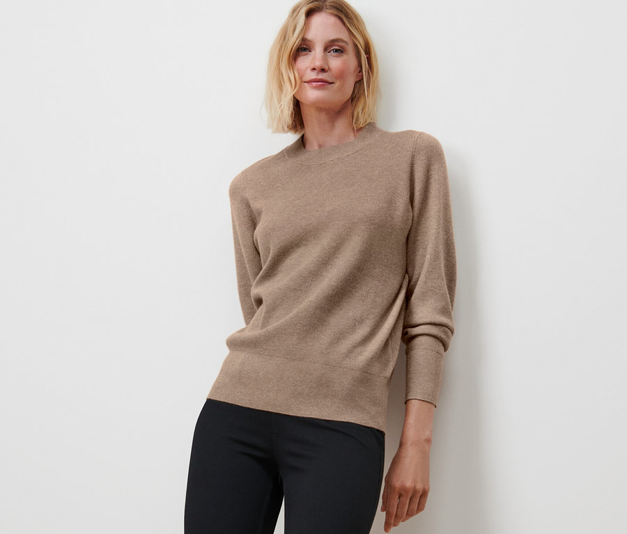 Frau posiert in Feinstrickpullover, beige meliert, und schwarzen Hosen.