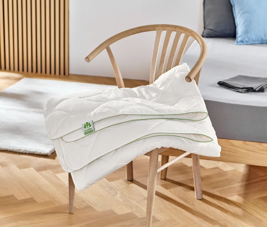 Ein gefaltetes, weißes irisette® greenline Baumwoll-Steppbett liegt auf einem Holzstuhl.