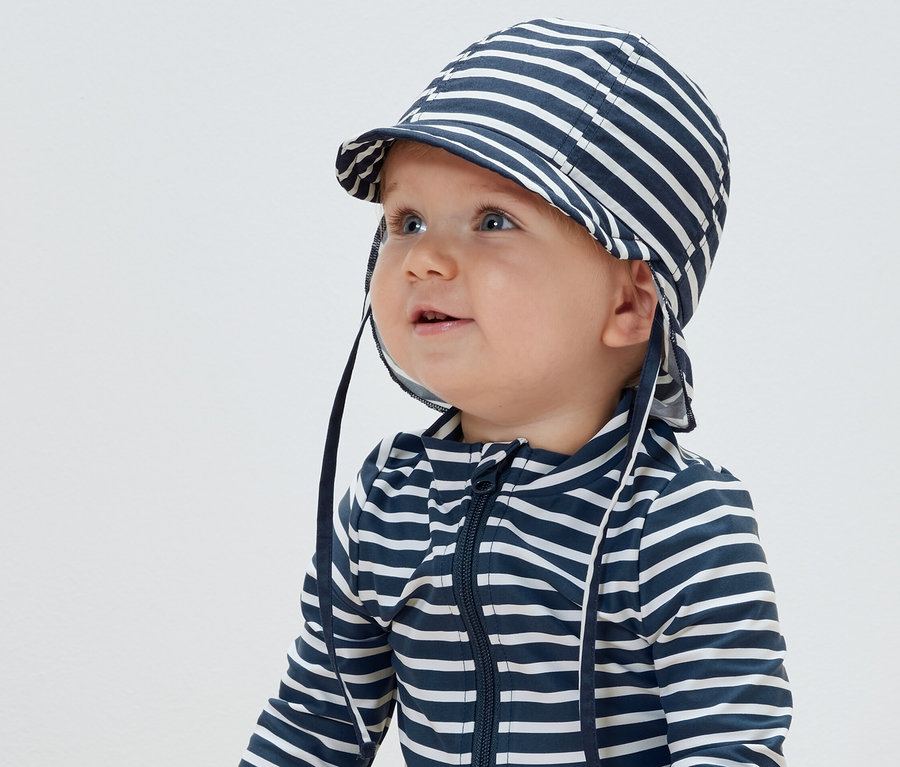 Baby trägt eine gestreifte Sonnenmütze mit UV-Schutzfaktor 80 und einen gestreiften Overall.