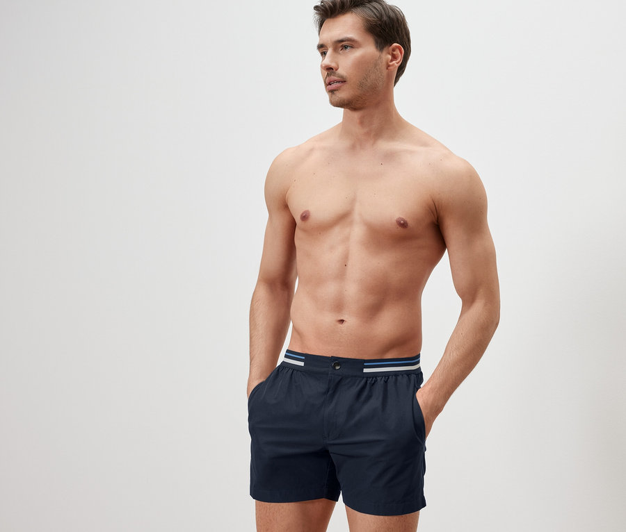 Mann mit Händen in den Taschen der dunkelblauen Badeshorts.