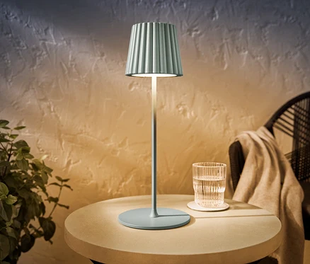 Eine salbeifarbene LED-Akku-Outdoor-Tischleuchte steht auf einem runden Tisch neben einem Glas Wasser.