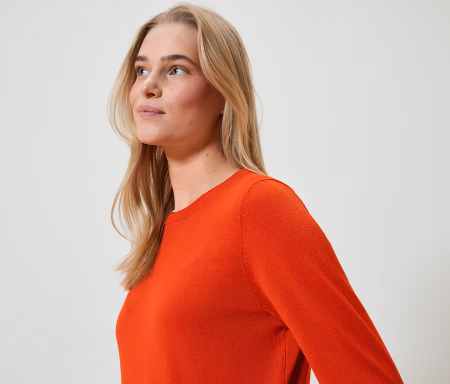 Porträt einer Frau mit blonden Haaren in einem orangefarbenen Pullover, die nach links schaut.