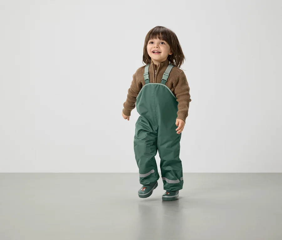 Lächelndes Kind mit braunen Haaren geht in grüner Latzhose und braunem Pullover.