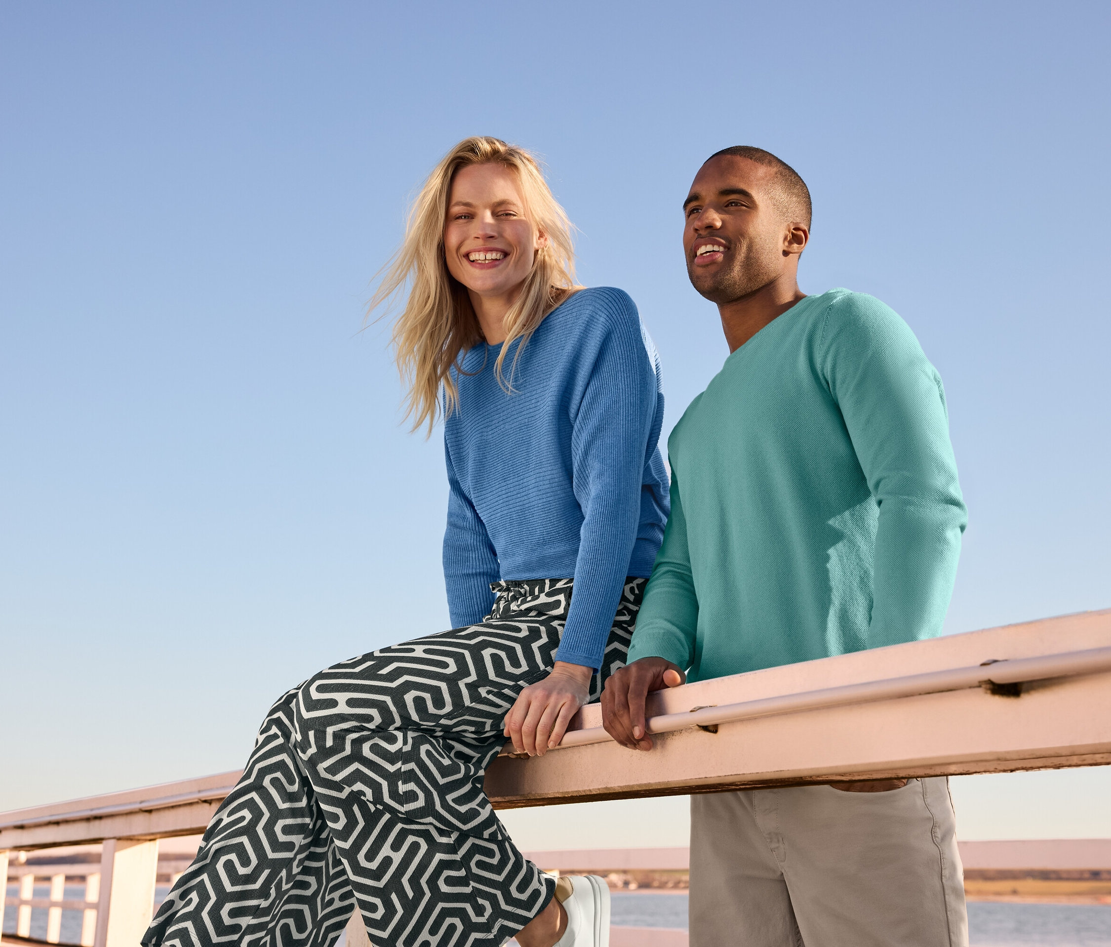 Eine Frau sitzt auf einem Geländer, trägt einen blauen Feinstrickpullover und eine bedruckte Schlupfhose in Crinkle-Optik. Daneben steht ein Mann in einem Strick-Pullover und einer 5-Pocket-Hose.