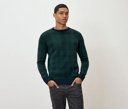 Mann posiert in grün kariertem Pullover und grauen Jeans.