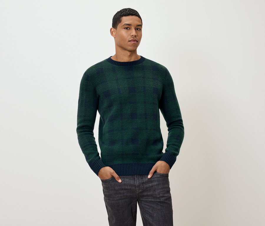 Mann posiert in grün kariertem Pullover und grauen Jeans.