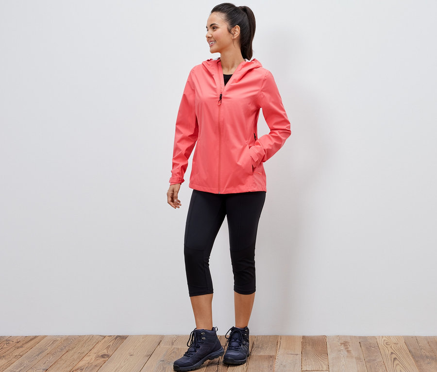 Eine lächelnde Frau posiert in einer korallenroten 2,5-Lagen-Outdoorjacke, einer schwarzen Dreiviertel-Leggings und Wanderschuhen.