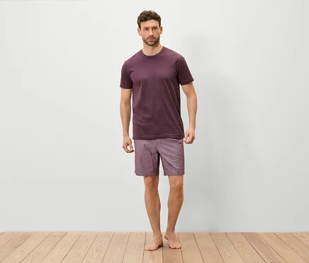 Mann steht barfuß auf einem Holzboden und trägt ein weinrotes T-Shirt und lila Shorts.