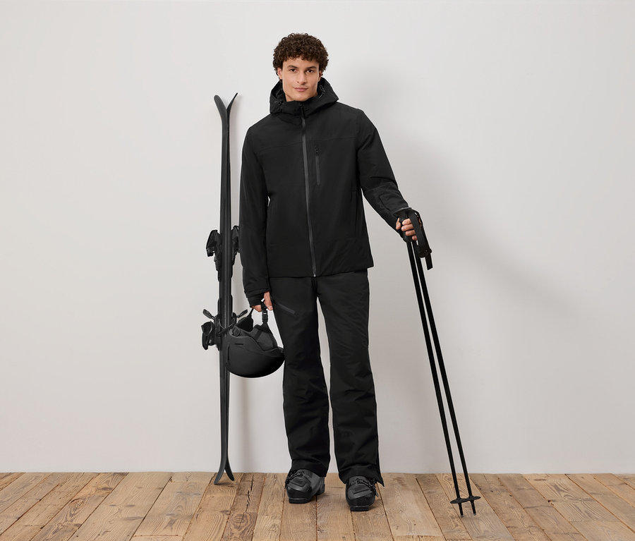 Mann in schwarzer Skibekleidung hält Stöcke, daneben Ski und Helm.