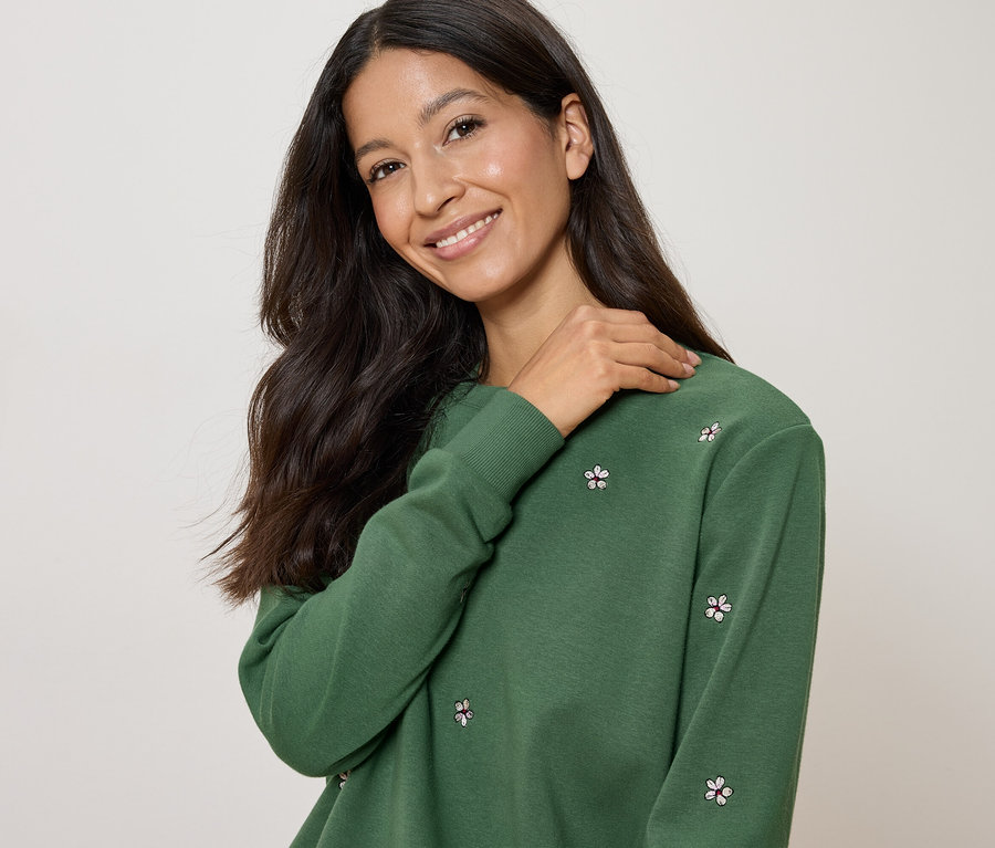 Frau mit dunklen, langen Haaren trägt ein grünes Sweatshirt mit Blumenmuster und legt ihre Hand auf ihre Schulter.