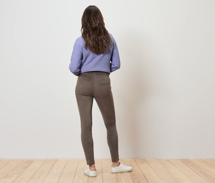 Eine Frau mit braunem, gewelltem Haar steht mit dem Rücken zum Betrachter und trägt ein lila Sweatshirt, braune Hosen und weiße Turnschuhe.