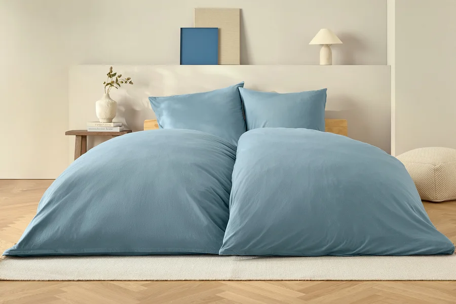Blaue Bettdecke und zwei Kissen auf einem Bett auf einem weißen Teppich.