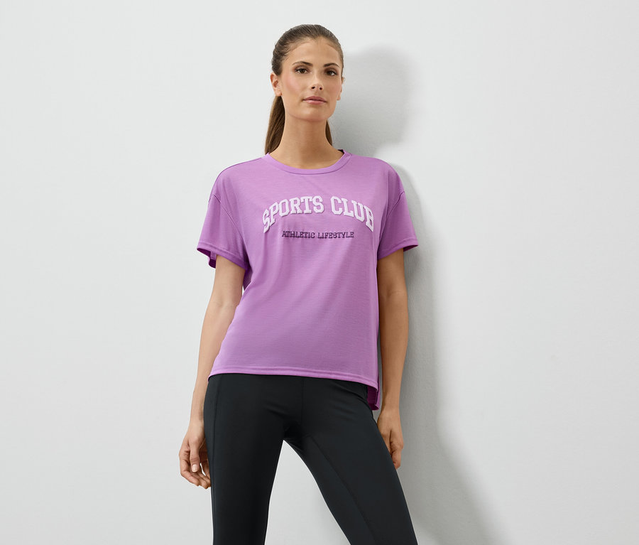 Frau in lila T-Shirt mit Aufdruck und schwarzen Leggings posiert vor einer weißen Wand.