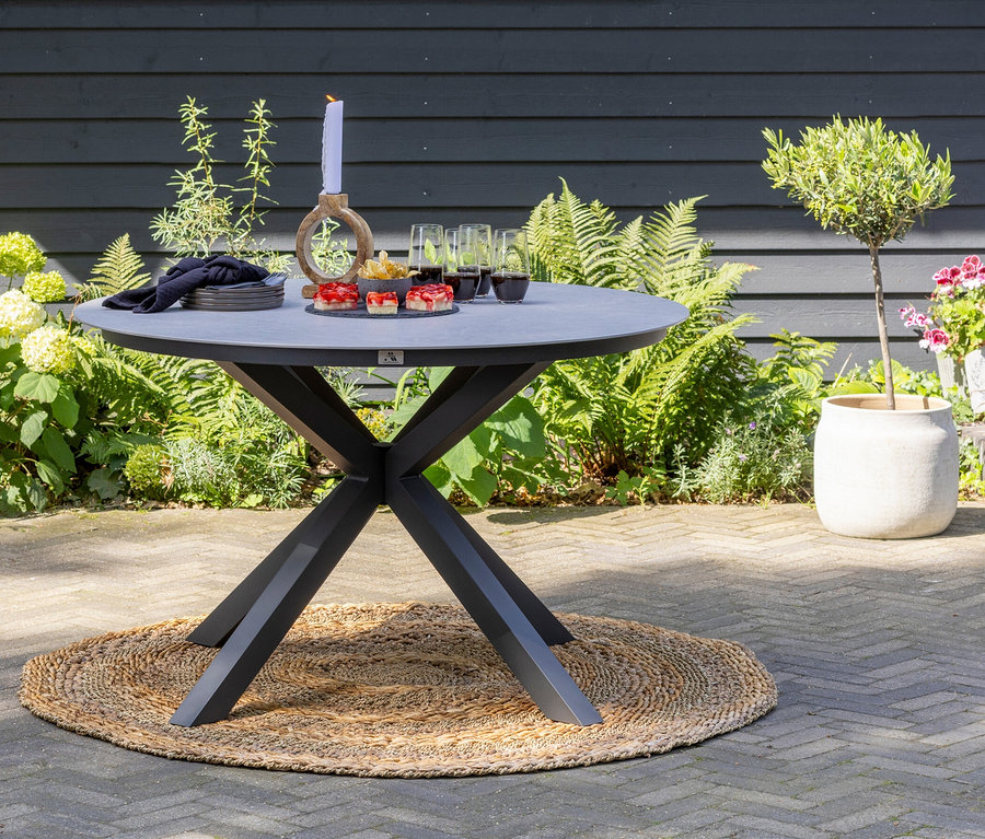 Außenszene mit einem runden deVries Tisch mit einem Durchmesser von ca. 120 cm, auf dem Speisen, Getränke und eine Kerze auf einem Jute-Teppich stehen.