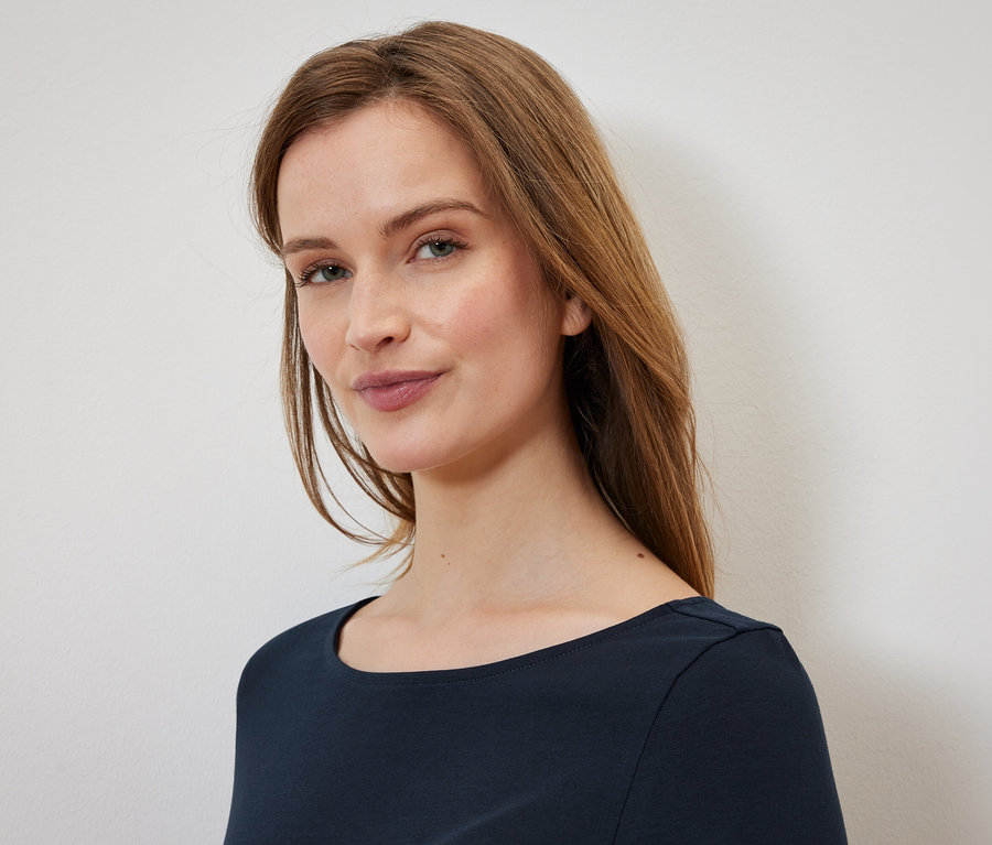 Porträt einer Frau mit langen, braunen Haaren und blauen Augen, die ein dunkelblaues T-Shirt trägt.