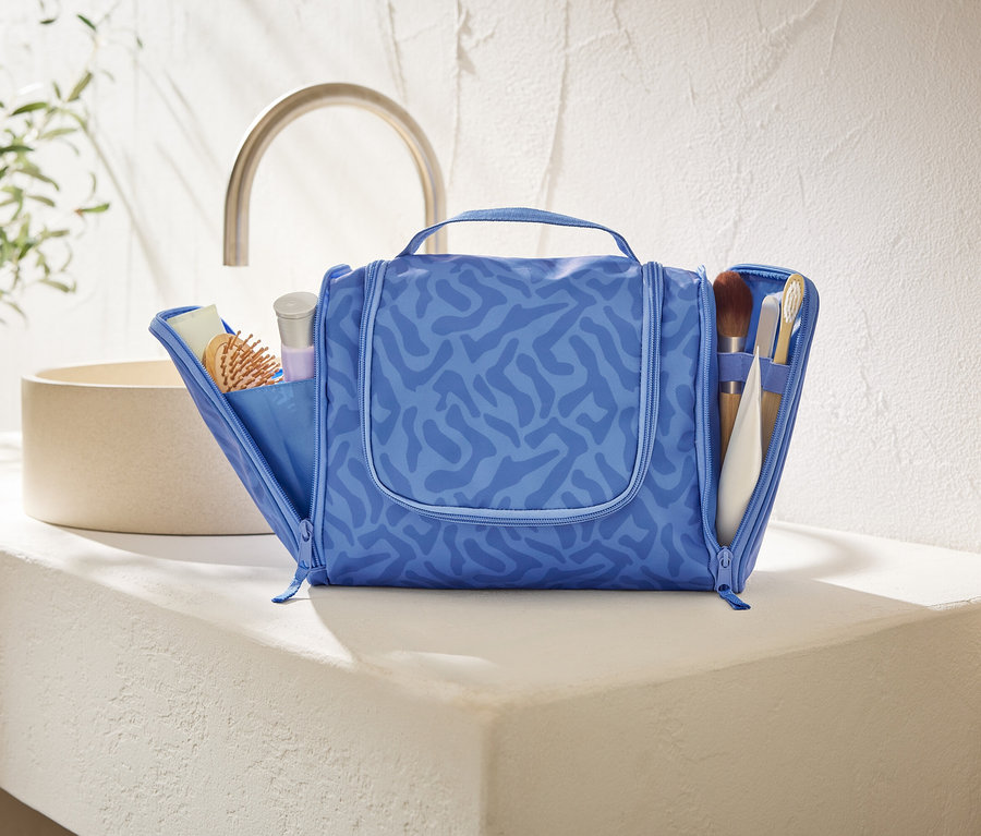 Eine offene, blaue Kosmetiktasche mit abstraktem Muster steht auf einer weißen Ablagefläche im Badezimmer.