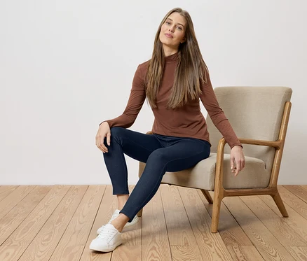 Frau mit braunem Oberteil und blauen Jeans sitzt auf einem Stuhl vor einer weißen Wand und einem Holzboden.