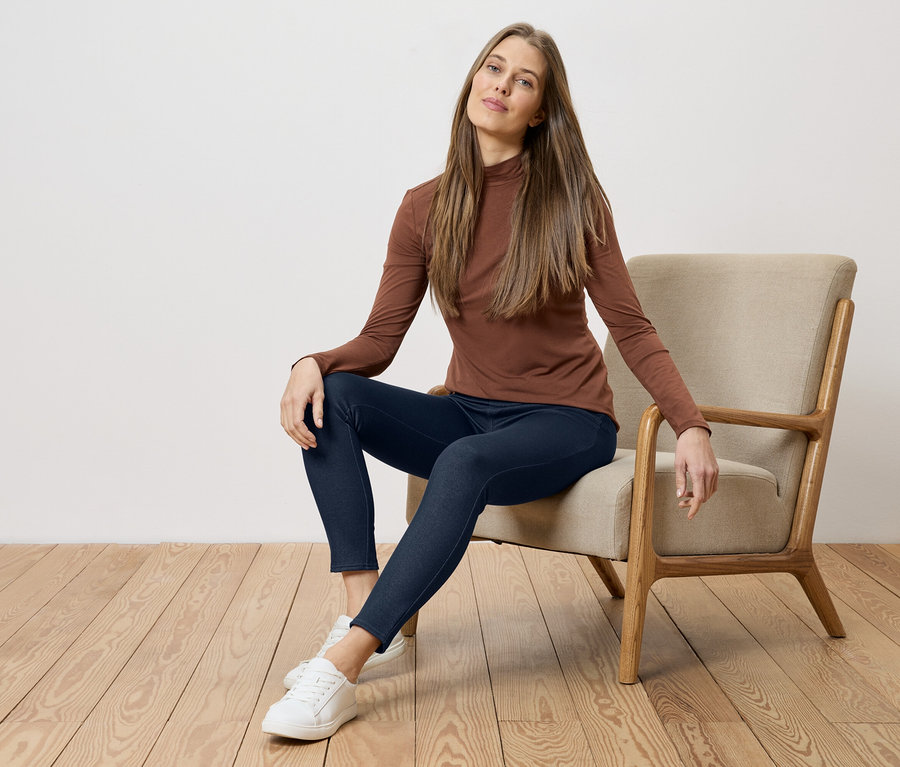 Frau mit braunem Oberteil und blauen Jeans sitzt auf einem Stuhl vor einer weißen Wand und einem Holzboden.