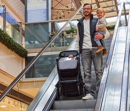 Mann mit Brille und Weste, der ein Kind auf dem Arm hält und einen Kinderwagen auf einer Rolltreppe in einem Einkaufszentrum schiebt.