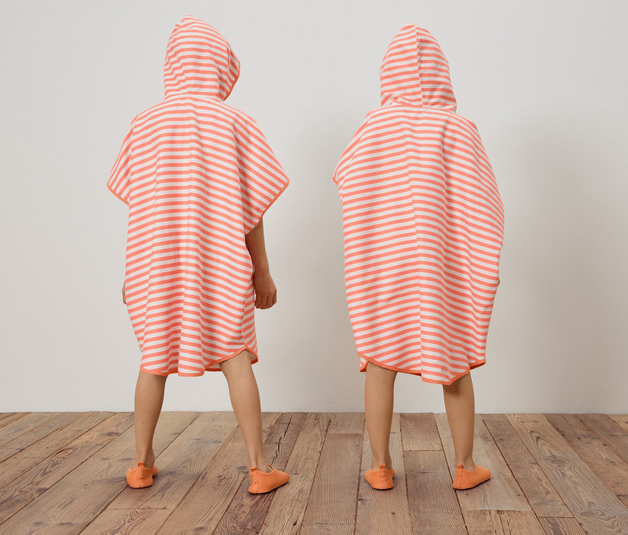 Zwei Kinder stehen mit dem Rücken zur Kamera und tragen gestreifte, korallenrote und weiße Ponchos mit Kapuze und passende Schuhe.